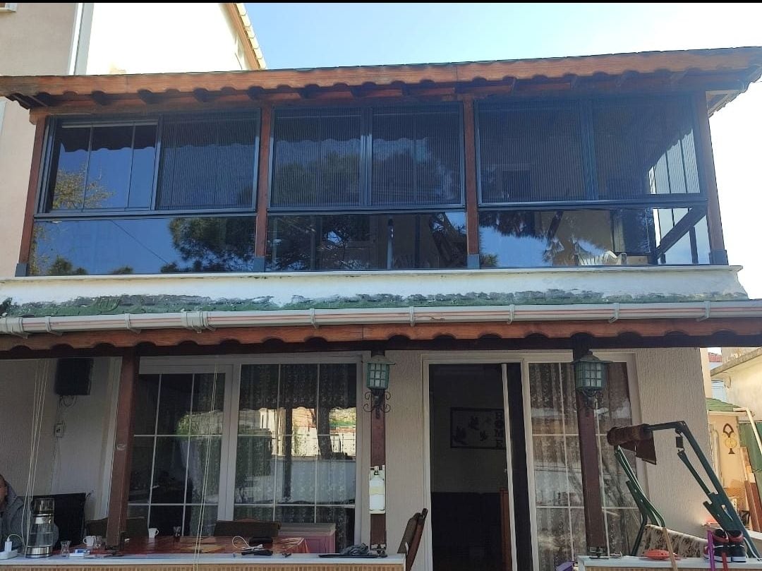 The exterior of a two-story house is visible, featuring large glass windows on the upper level that provide a clear view of the surroundings. A shaded porch is seen at the front, with outdoor furniture and potted plants adding to the inviting appearance.
