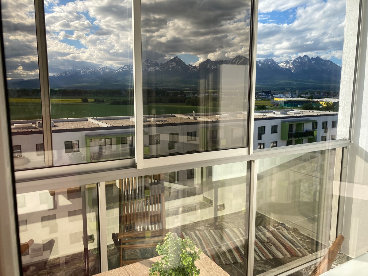 A view through large glass windows reveals a scenic panorama of the High Tatras, with mountains capped in snow. A wooden chair on a balcony invites relaxation, while green fields extend below, reflecting a serene atmosphere.