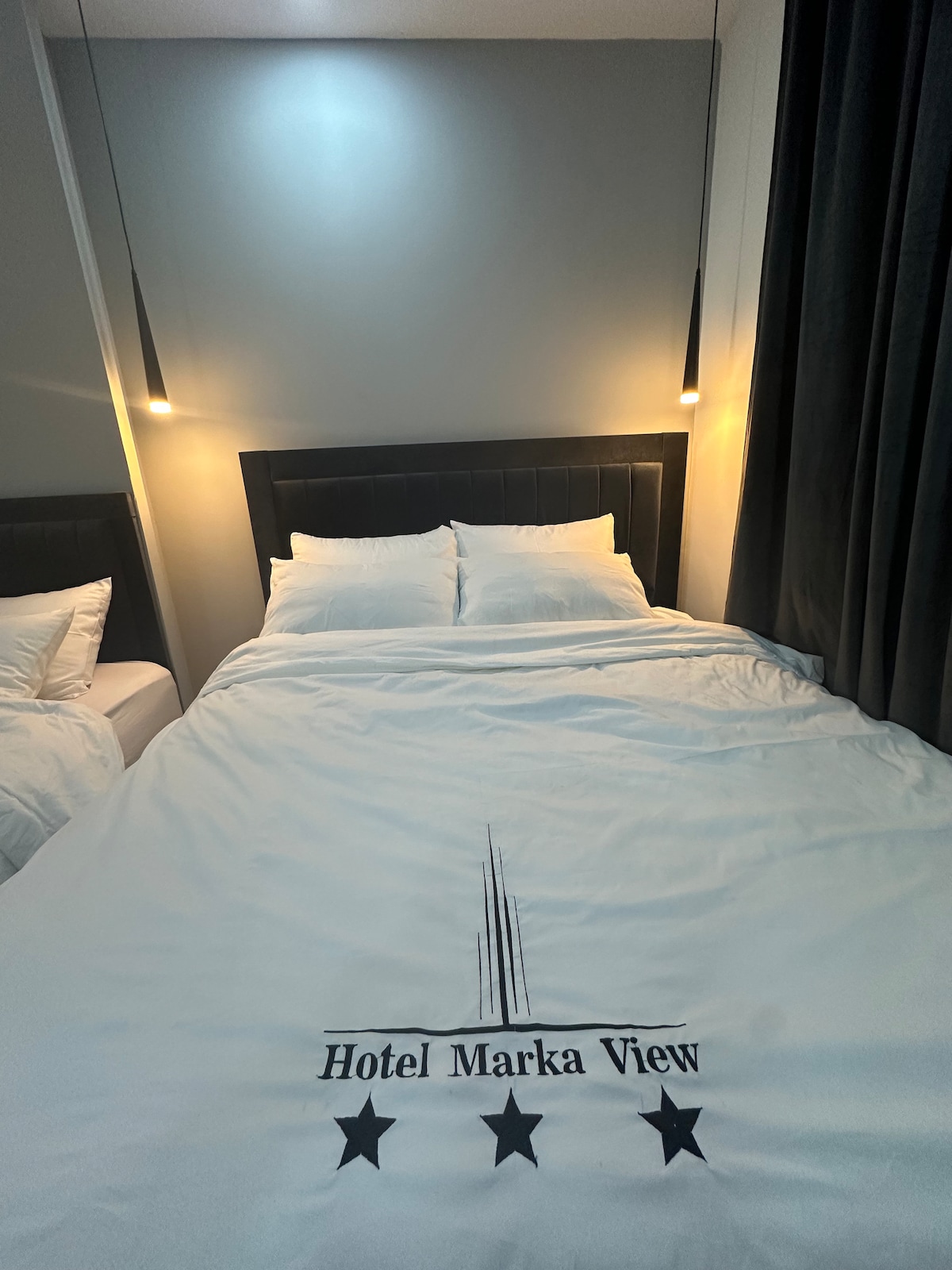 A cozy sleeping area features a spacious bed dressed in a white duvet with the Hotel Marka View logo. Soft lighting is provided by pendant lights above the bed, illuminating the neutral walls and creating a serene environment. Dark curtains frame a side window.