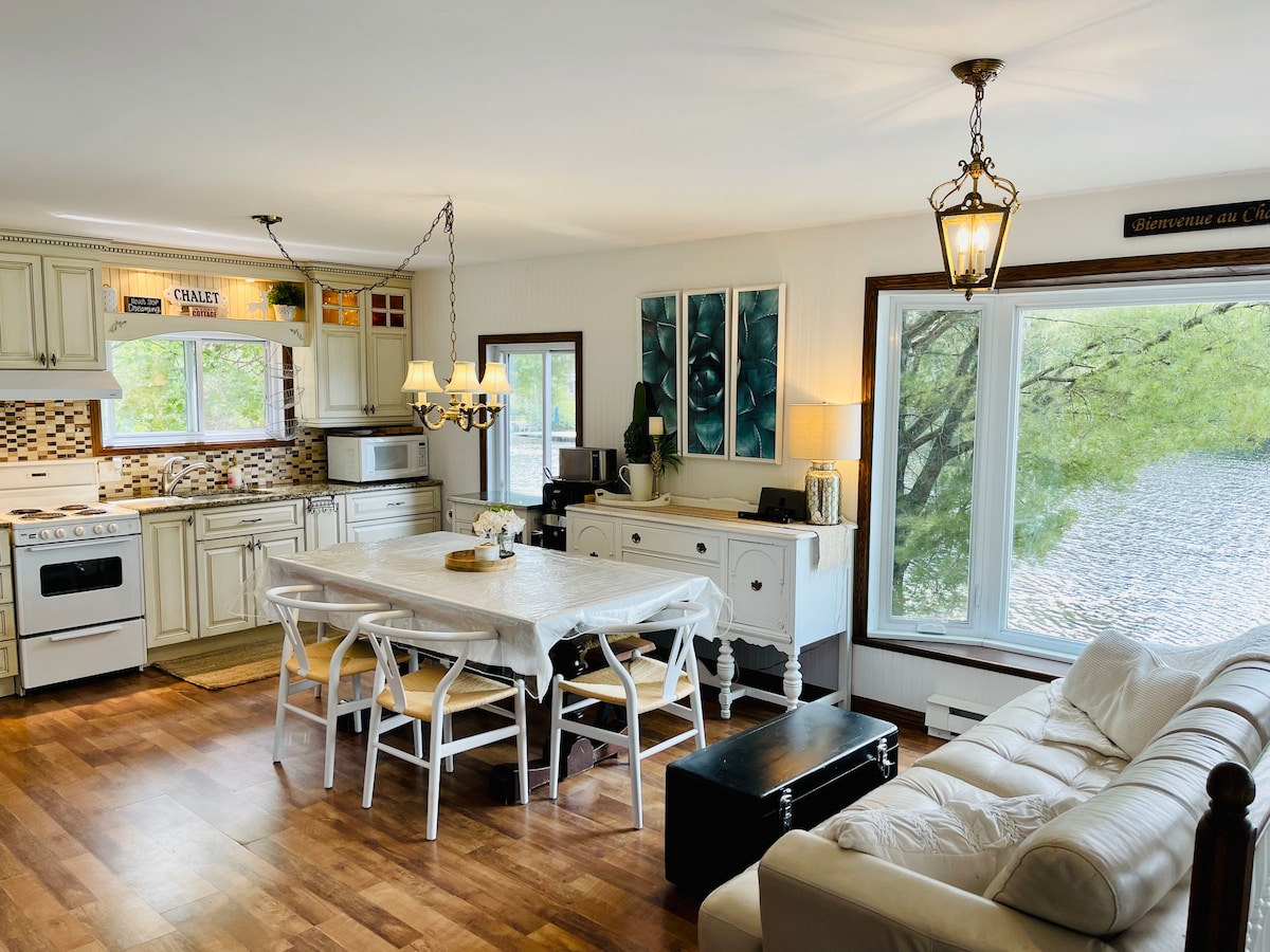 A well-equipped kitchen and dining area is showcased, featuring a large table surrounded by white chairs. Natural light streams through the windows, which offer views of the lake. A cozy living space is visible, with a light-colored sofa and decorative elements enhancing the ambiance.