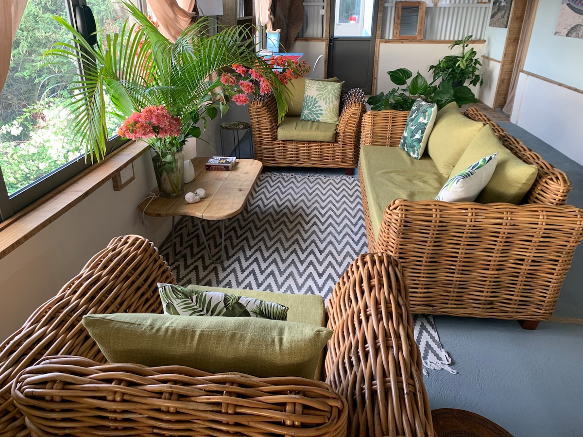 A relaxed seating area features wicker furniture adorned with green and patterned cushions. A natural wood coffee table sits in the center, surrounded by various indoor plants and vibrant flowers, creating a fresh and inviting atmosphere.