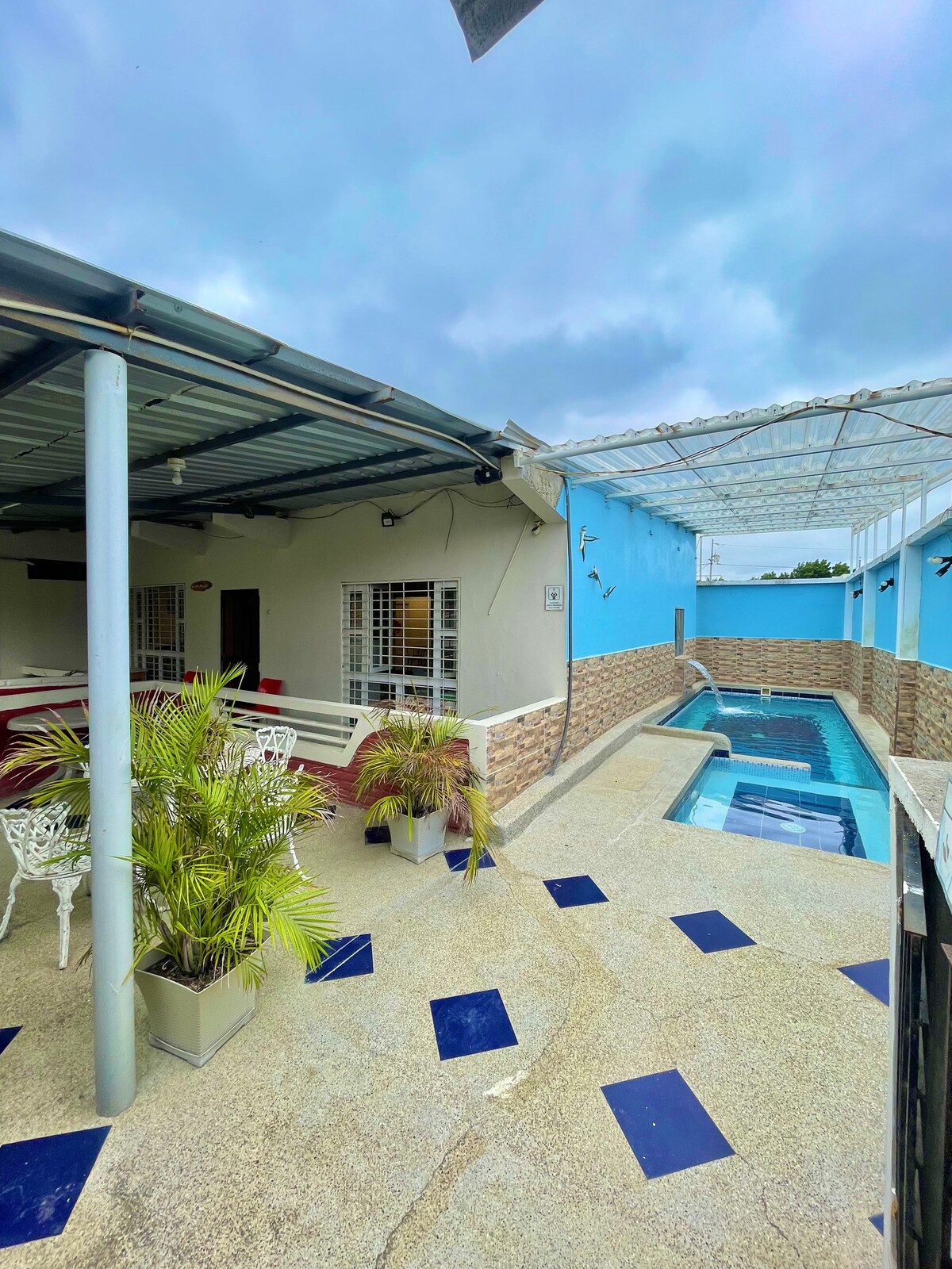 An outdoor area showcases a partially covered patio with white seating and potted plants. A narrow swimming pool runs alongside the wall, with light blue painted walls adding a refreshing touch. The floor is adorned with blue and gray tiles, creating a cohesive look.