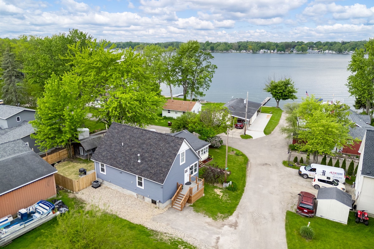 An aerial view captures Maddison House on Grace, showcasing its modern exterior and proximity to a serene lake. Lush green trees frame the backyard, while the nearby road leads to additional homes. The peaceful waters reflect the sky, enhancing the tranquil setting.