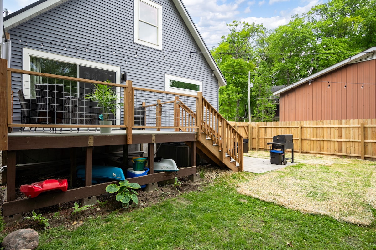 The outdoor area features a wooden deck with steps leading down to a grassy yard bordered by a wooden fence. A small grill and seating are visible on the deck, along with colorful outdoor toys stored nearby. The setting is well-maintained with green grass and a manicured appearance.