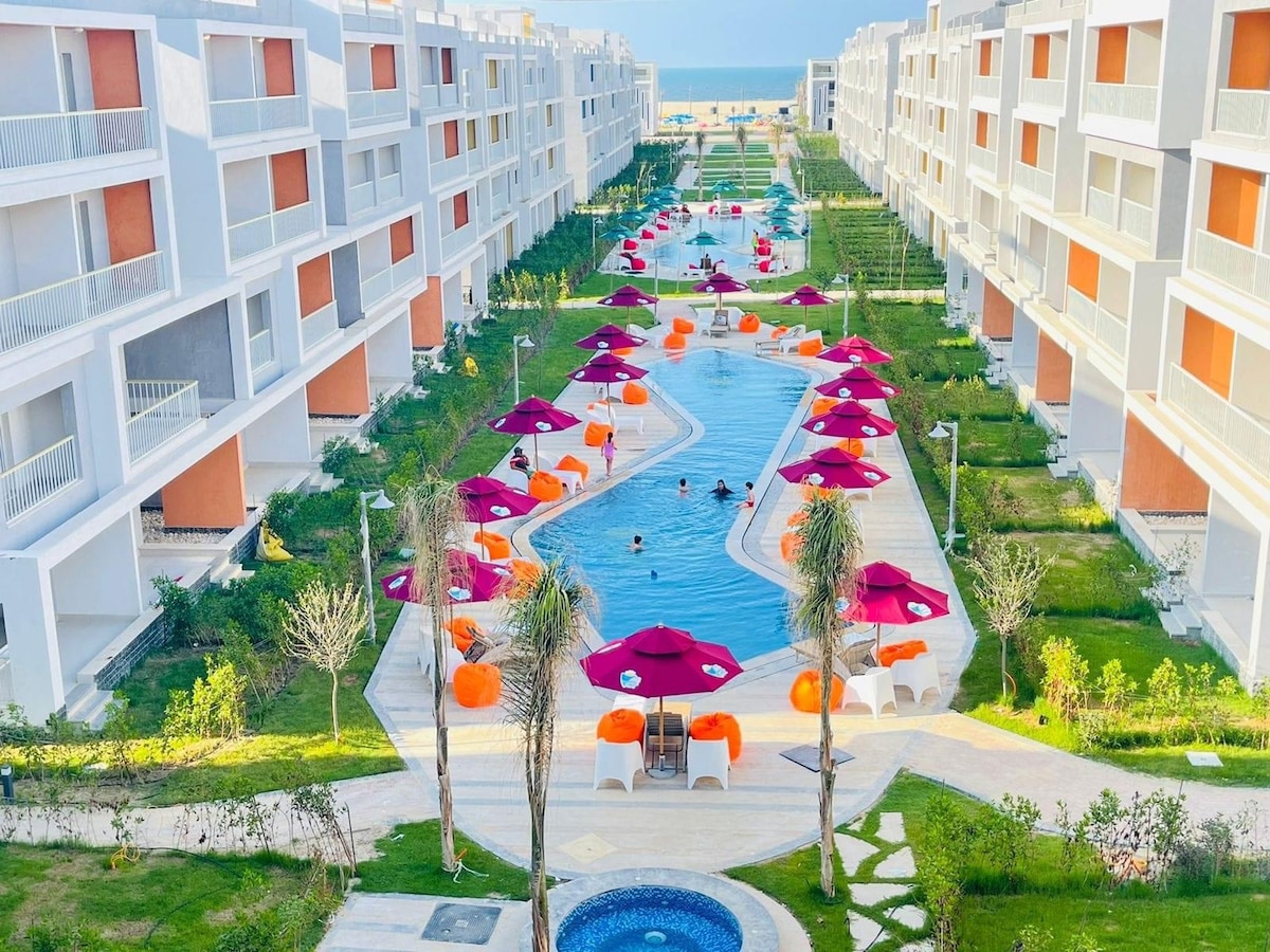 The image captures a spacious outdoor area featuring a winding pool set among greenery. Colorful lounge chairs and umbrellas are arranged along the poolside, providing shaded spots for relaxation. Light reflects off the water, enhancing the inviting ambiance of the resort.