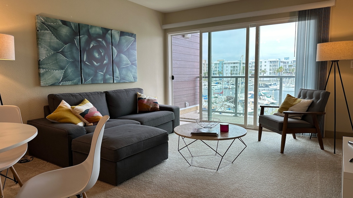 A comfortable living area features a spacious gray sofa paired with an accent chair. A modern coffee table is positioned in front, and soft throw pillows add color. Large sliding glass doors offer a view of the marina, allowing natural light to fill the room.
