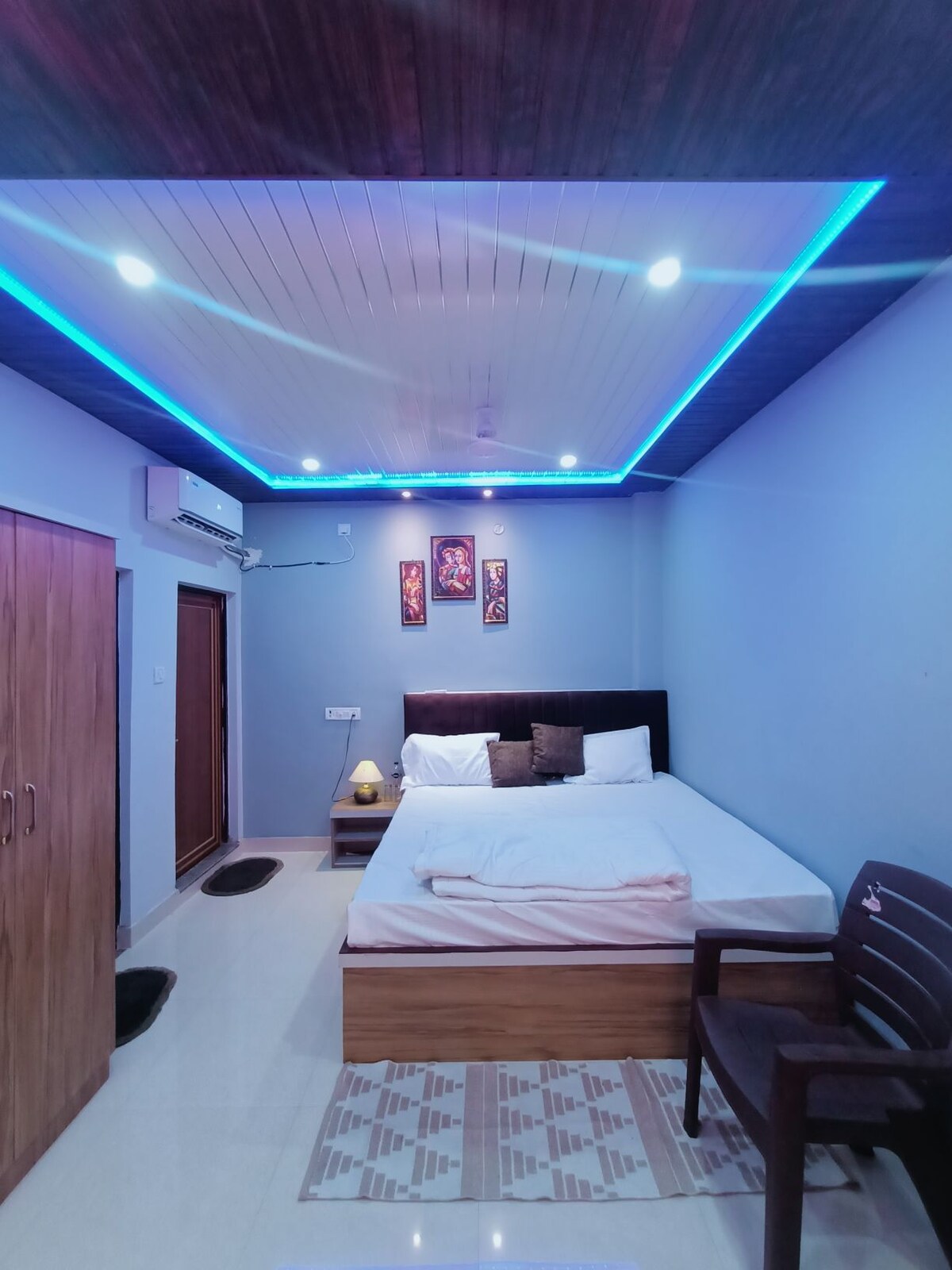 A cozy sleeping area features a spacious bed with white linens against a backdrop of calming blue walls. Soft lighting from LED strips above creates an ambient glow. A wooden chair and a small table are placed beside the bed, with a decorative rug adding warmth to the floor.