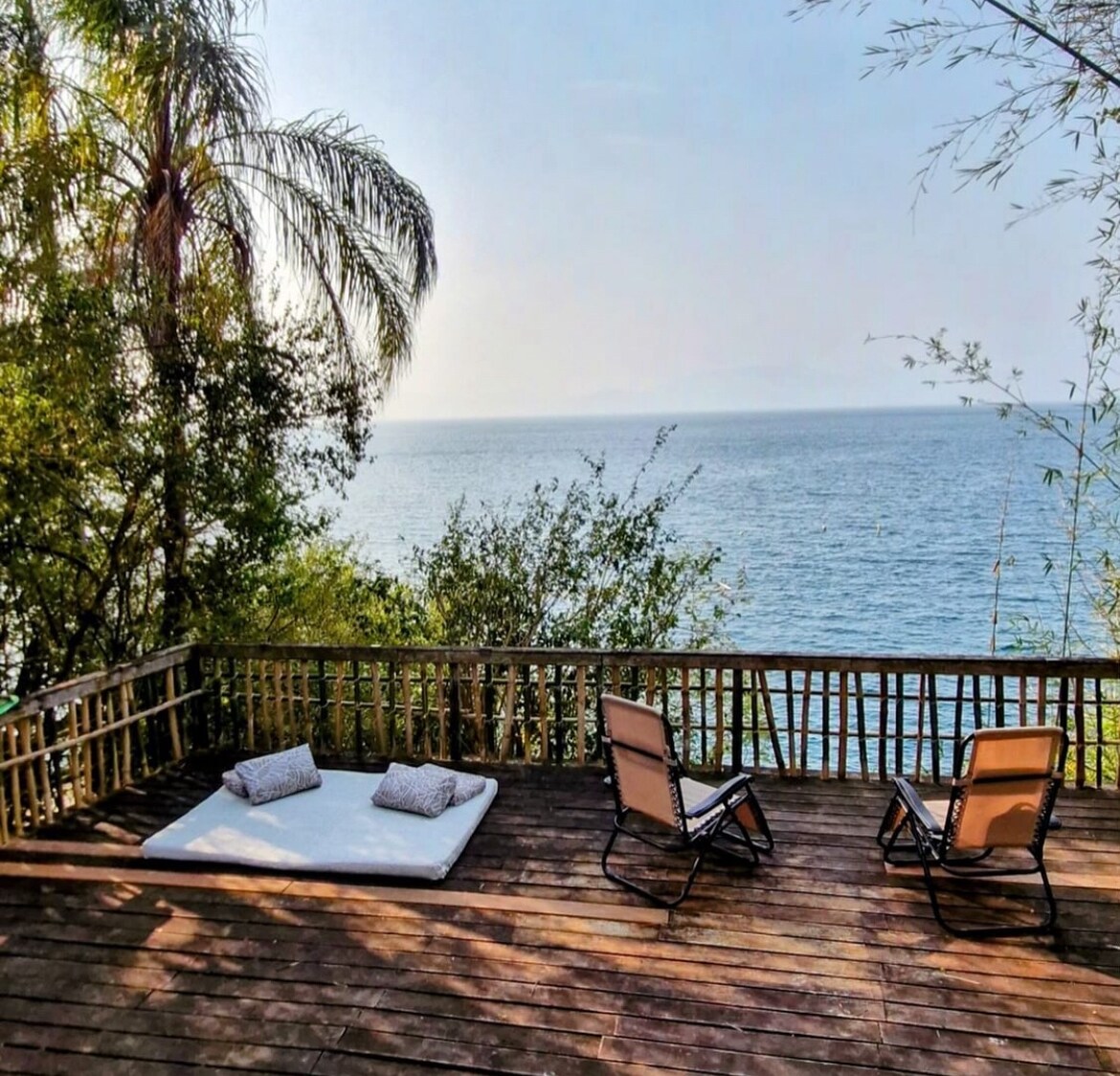 A spacious wooden deck overlooks serene waters, blending with lush greenery. Two lounge chairs are positioned near a cushioned area for relaxation. The distant horizon features a calm, reflective surface under a clear sky.