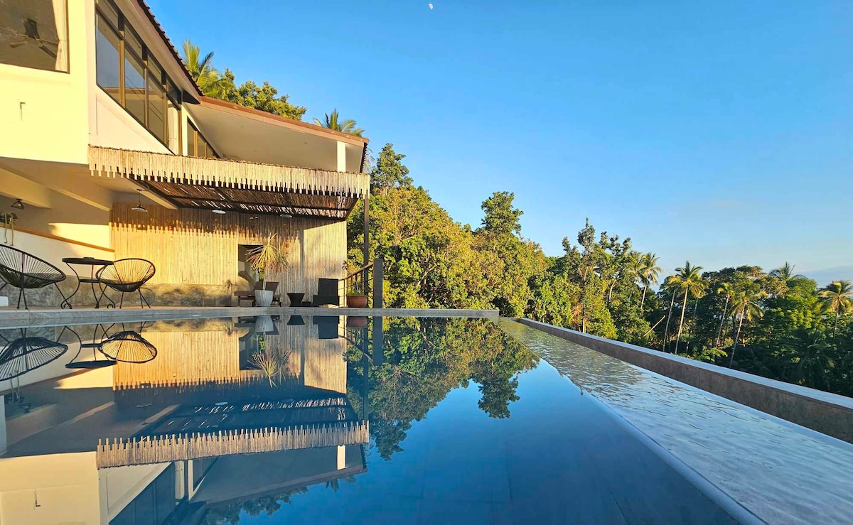 The image captures a serene pool area with a clear, reflective surface, showcasing the surrounding tropical landscape. Lush greenery and palm trees are visible in the background, while the modern structure is partially seen, featuring a terrace with lounge seating.