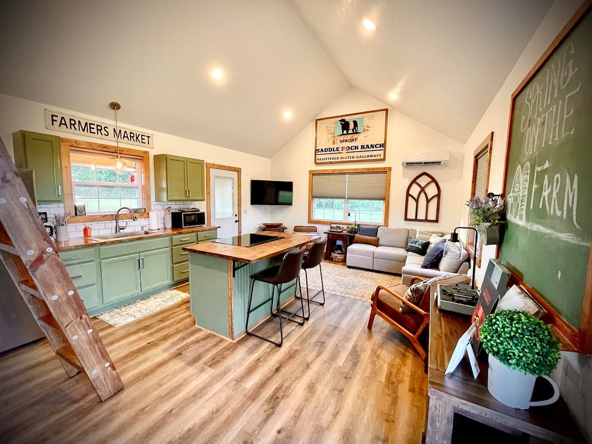 The open kitchen and common area feature a spacious layout with vaulted ceilings. Green cabinetry complements the wood accents. A sectional couch, dining tables, and large windows offer natural light, creating a welcoming space. A decorative sign adds a rustic charm to the atmosphere.
