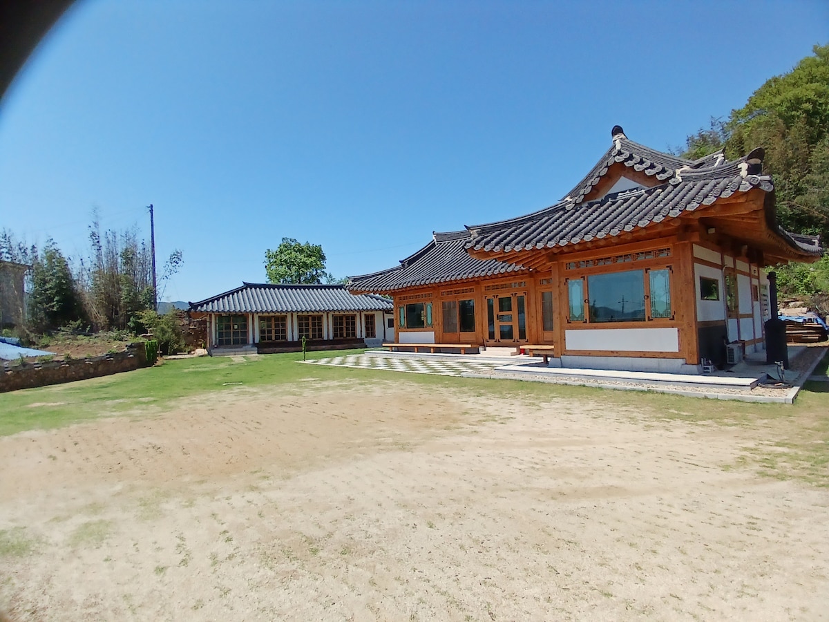 A traditional Korean hanok is depicted, featuring distinct curved roofs with tiles and wooden accents. The structure is complemented by spacious outdoor areas, including a manicured lawn and pathways leading to additional buildings. Natural surroundings contribute to a serene environment.
