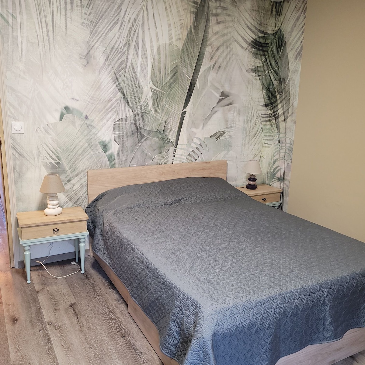 A spacious bedroom features a bed dressed in a grey quilt, flanked by two nightstands with table lamps. The wall behind displays a tropical leaf motif, while the floor is made of light wooden planks, creating a serene environment.