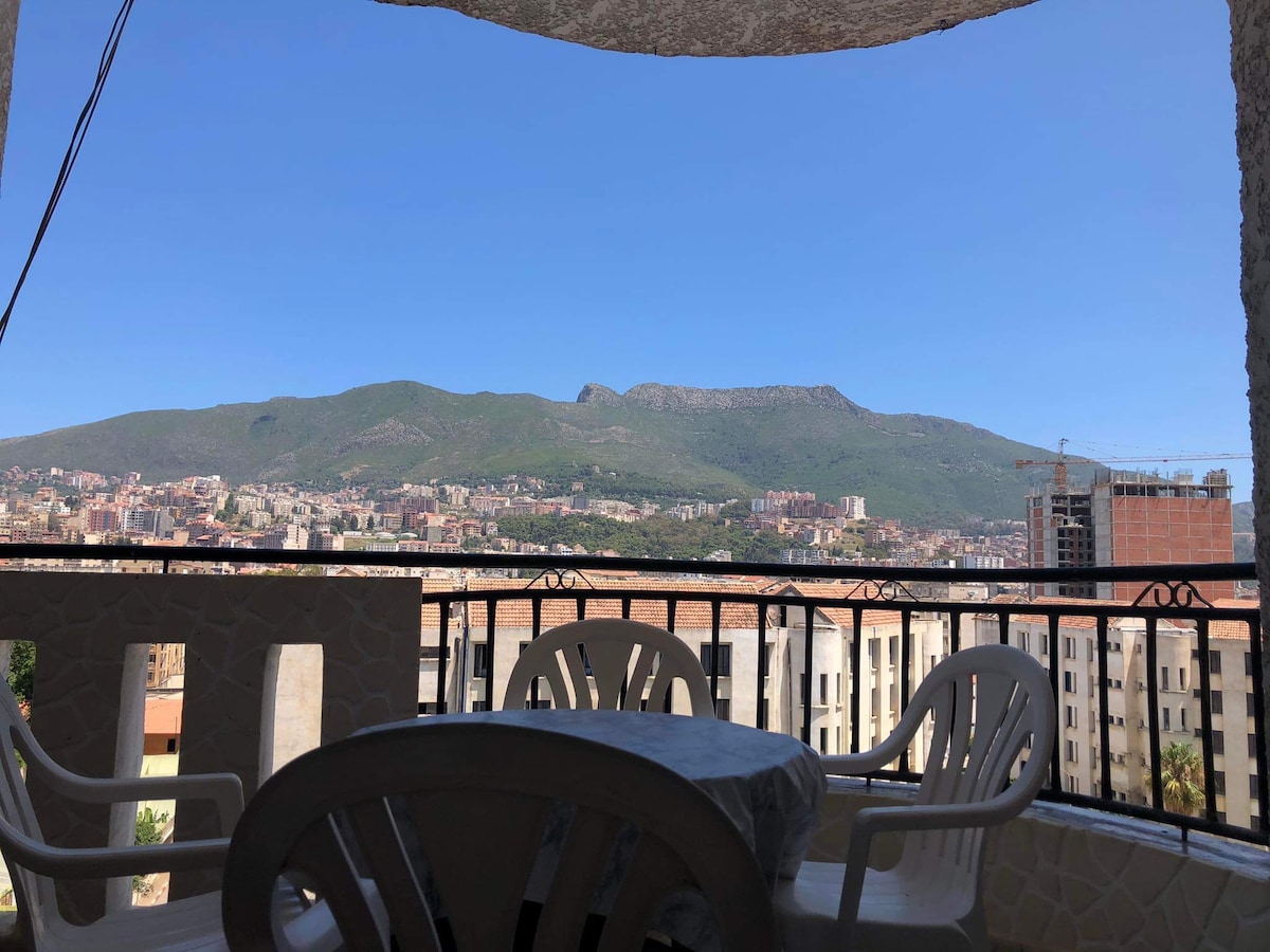 A balcony view reveals a panoramic landscape of rolling mountains under a clear blue sky. The image includes a table and several white chairs, offering a comfortable space to enjoy the scenery.
