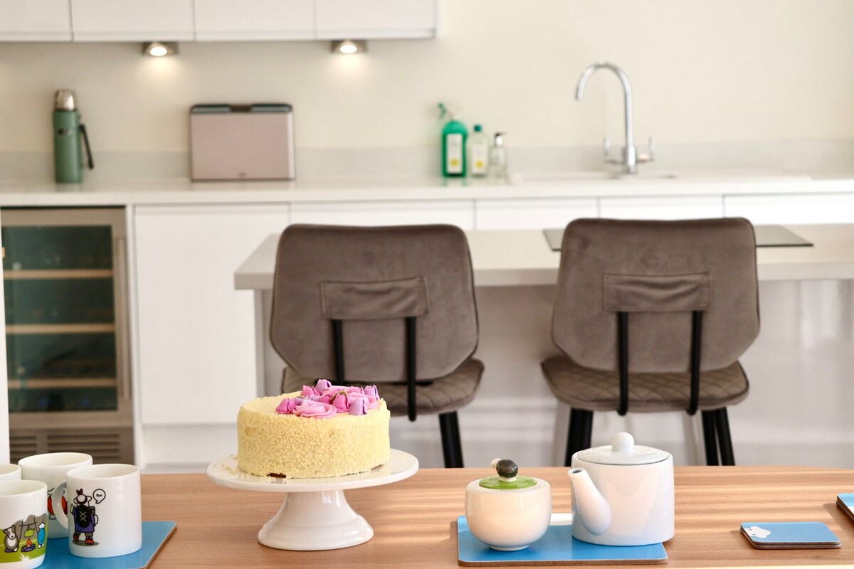 A modern kitchen area is showcased, featuring a light-colored countertop and sleek cabinetry. Two gray high-backed stools are positioned at the kitchen island. A cake with pink frosting sits on a stand, surrounded by a teapot and cups, indicating a welcoming space for gatherings.