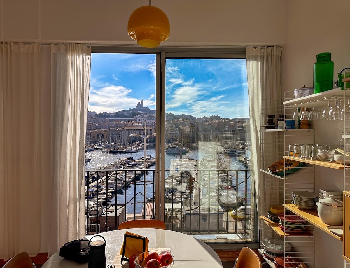 A dining area is presented with a circular table and access to an expansive view through large glass doors. The scene outside features the vibrant port with boats and the silhouette of a historic landmark in the background. Shelves filled with dishware are visible to the side.