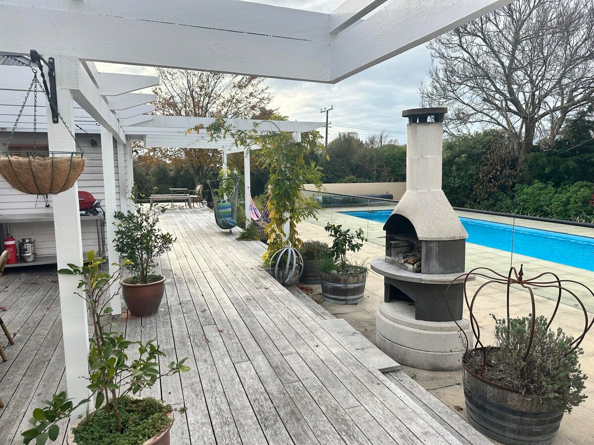 A wooden deck surrounds a large pool, with various potted plants placed thoughtfully. A brick barbecue structure is positioned next to the pool, and a shaded area features an inviting seating arrangement under a white pergola. Lush greenery borders the space.