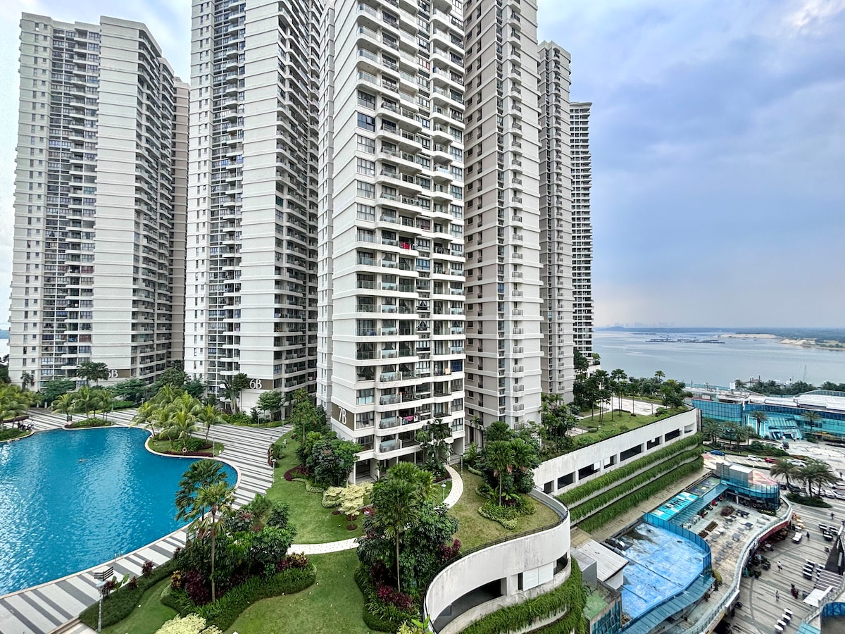The image showcases a sprawling residential complex surrounded by lush landscaping. A large swimming pool reflects the sky, while multiple high-rise buildings form a modern backdrop. Views extend towards the water, offering a sense of tranquility in the setting.