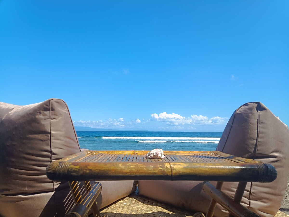 A natural bamboo table is positioned between two soft lounge cushions, offering a clear view of the ocean. Calm waves gently lap at the shore under a bright blue sky, creating a serene backdrop for relaxation.