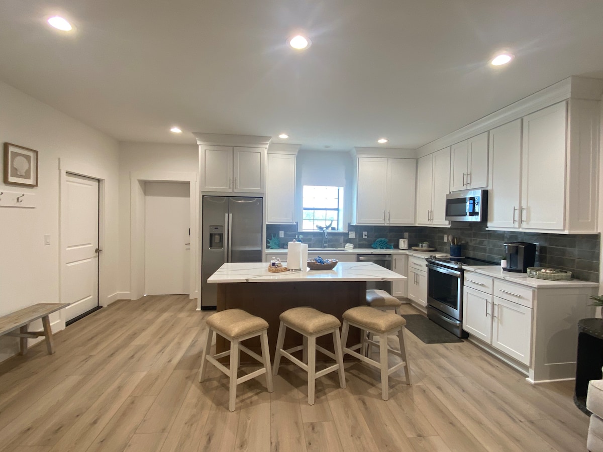 The modern kitchen features light cabinetry and a central island with seating for four. Stainless steel appliances are present, including a refrigerator and oven. Natural light streams through the window, highlighting the clean lines and neutral color palette of the space.