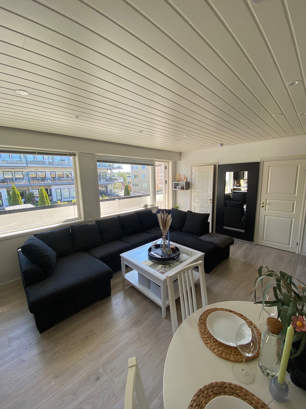 A spacious living area features a large sectional sofa positioned along the wall, with sunlight streaming through multiple windows. A white coffee table sits at the center, adorned with decorative items. A dining table appears nearby, set with woven place mats and glassware.