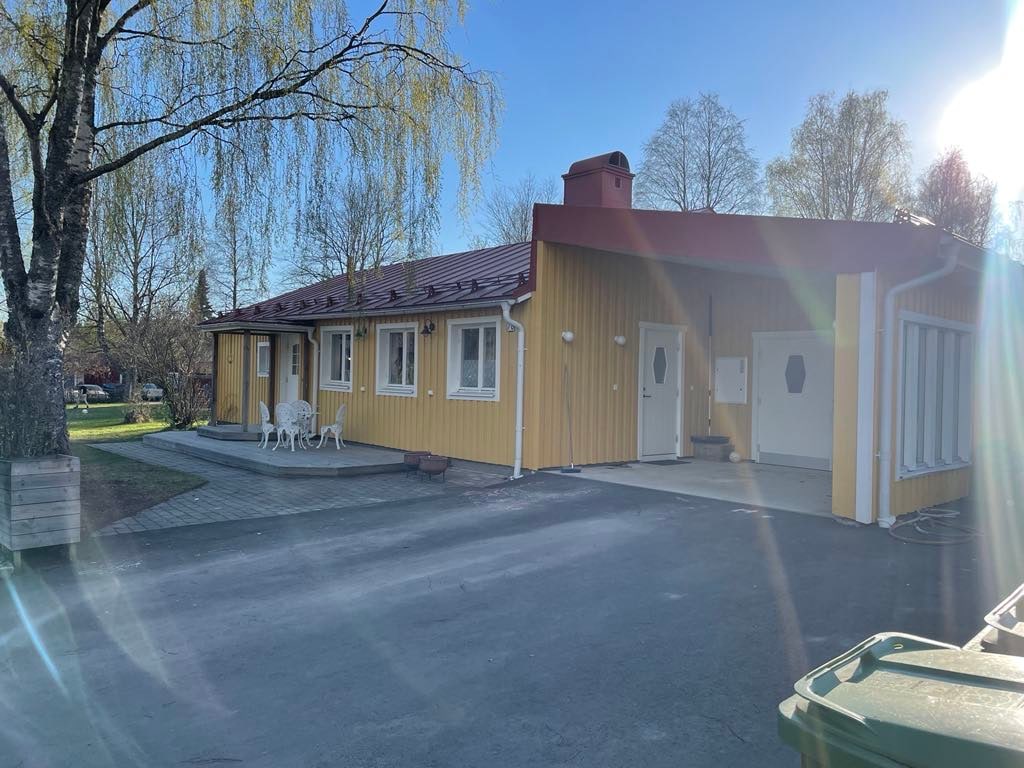 A yellow house with a red roof is positioned among trees and greenery. A small patio area features white outdoor chairs. The entrance displays two doors, with a paved walkway leading to the driveway. Natural light illuminates the scene, enhancing the bright exterior.