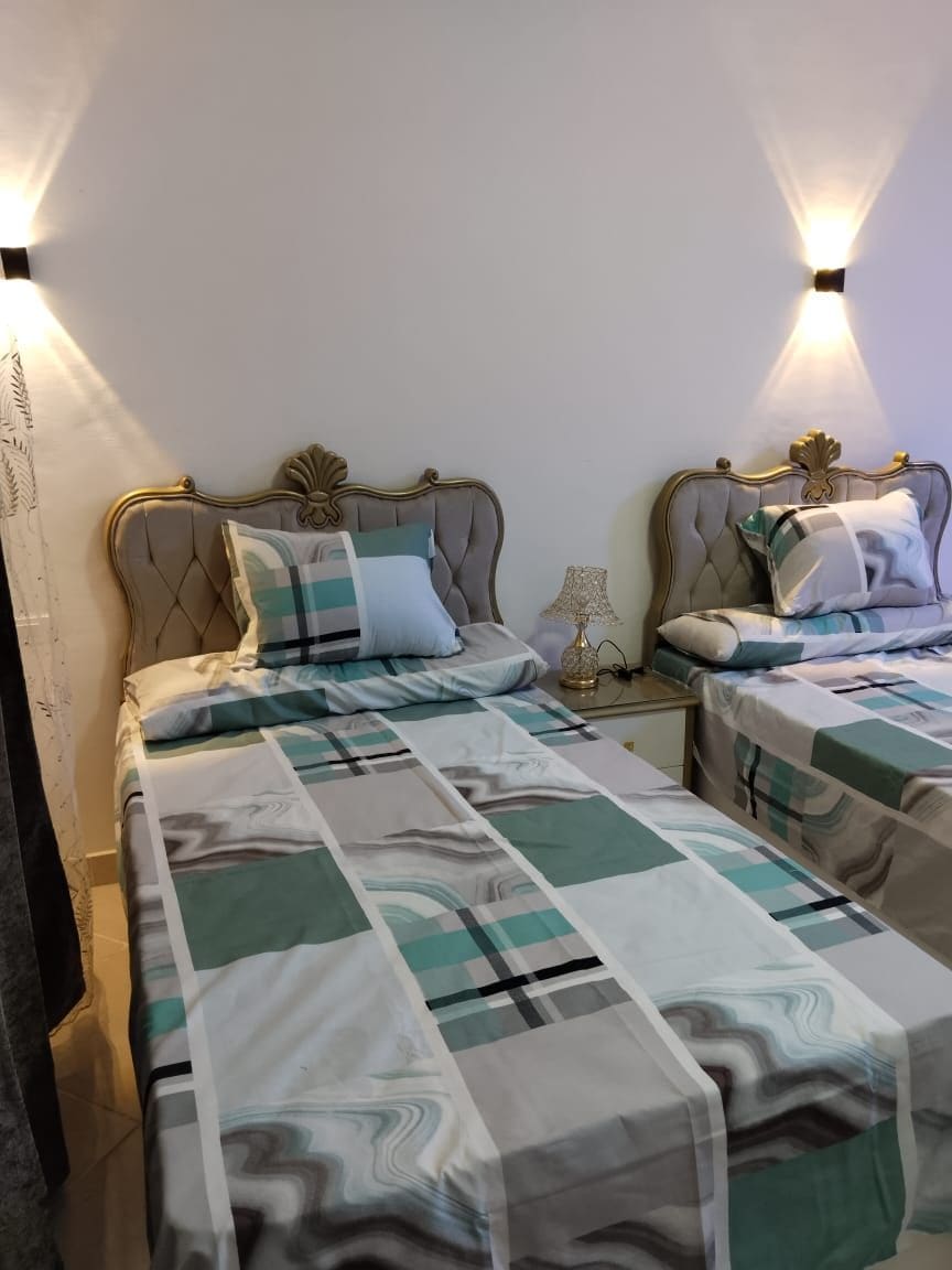 A well-appointed bedroom features two elegant beds with tufted headboards. Each bed is adorned with patterned bedding in soothing colors. Soft lighting from wall sconces enhances the calming atmosphere, while a small bedside table hosts a decorative lamp.