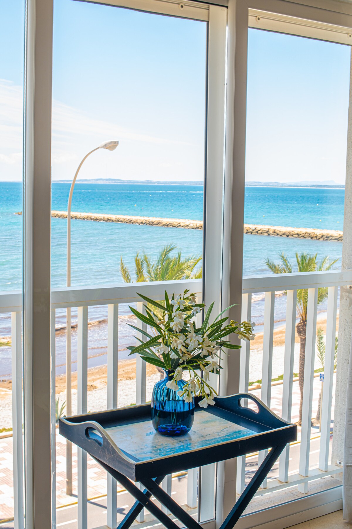 A table with a blue vase containing fresh white flowers is positioned in front of large glass windows. The expansive view overlooks the sea and beach, with palm trees visible along the shoreline and a clear sky in the background.