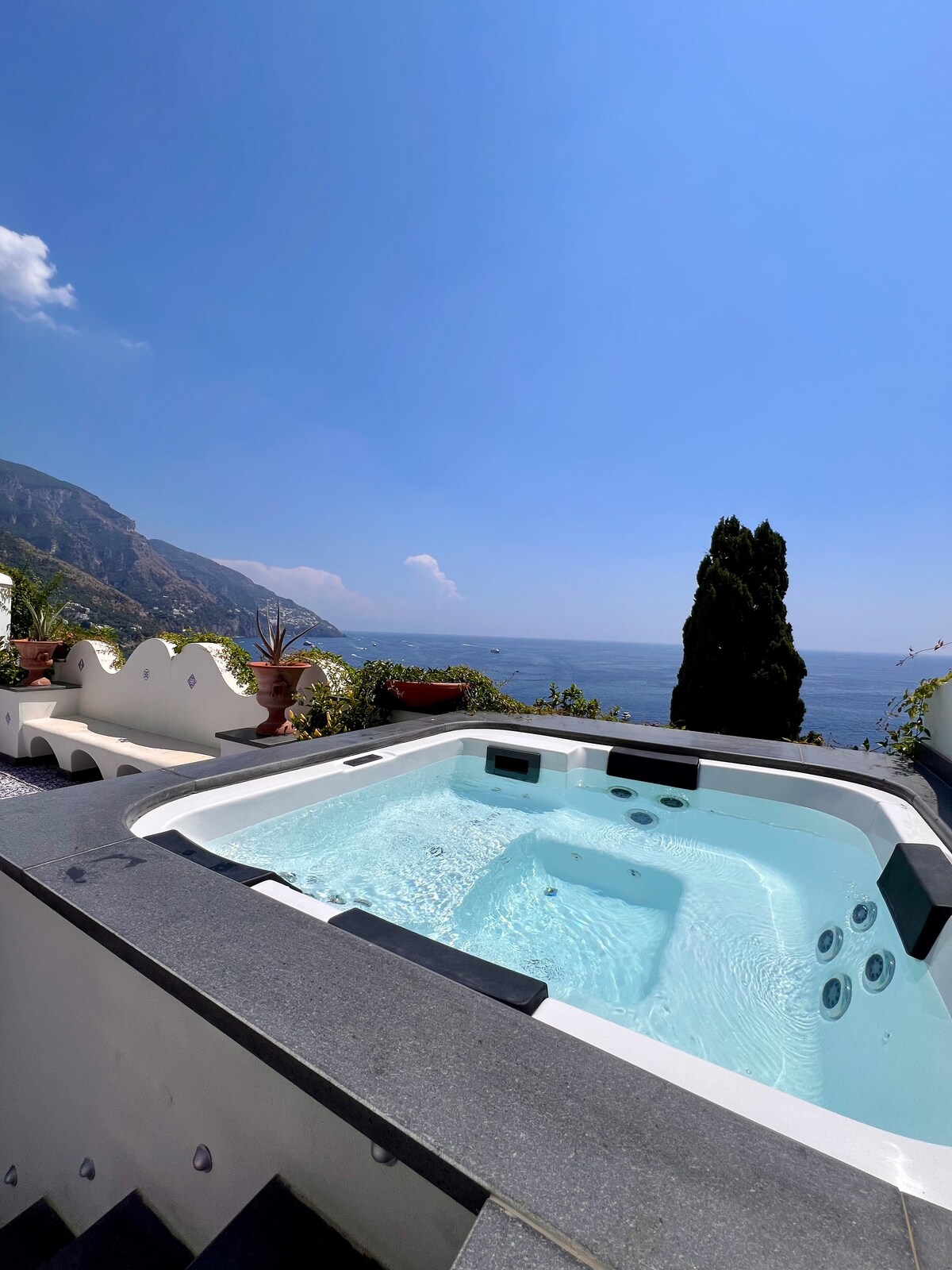 A spacious outdoor terrace features a jacuzzi surrounded by lush greenery and terracotta planters. The panoramic view of the sea extends towards the horizon, while the backdrop showcases gently sloping mountains and a bright blue sky.