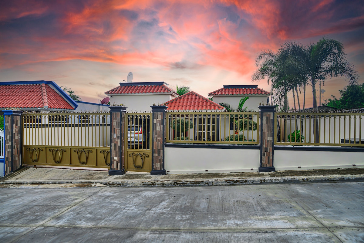 A spacious property is enclosed by a decorative fence featuring golden accents. The vibrant sunset casts warm hues over the roof tiles of the home, surrounded by tropical greenery. An inviting ambiance is conveyed by the overall setting.