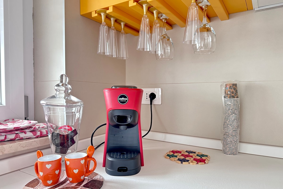 A modern kitchen corner features a red coffee machine next to two orange cups. Above, shelves hold glasses, while a decorative jar and a stack of colorful coasters add visual interest to the countertop.