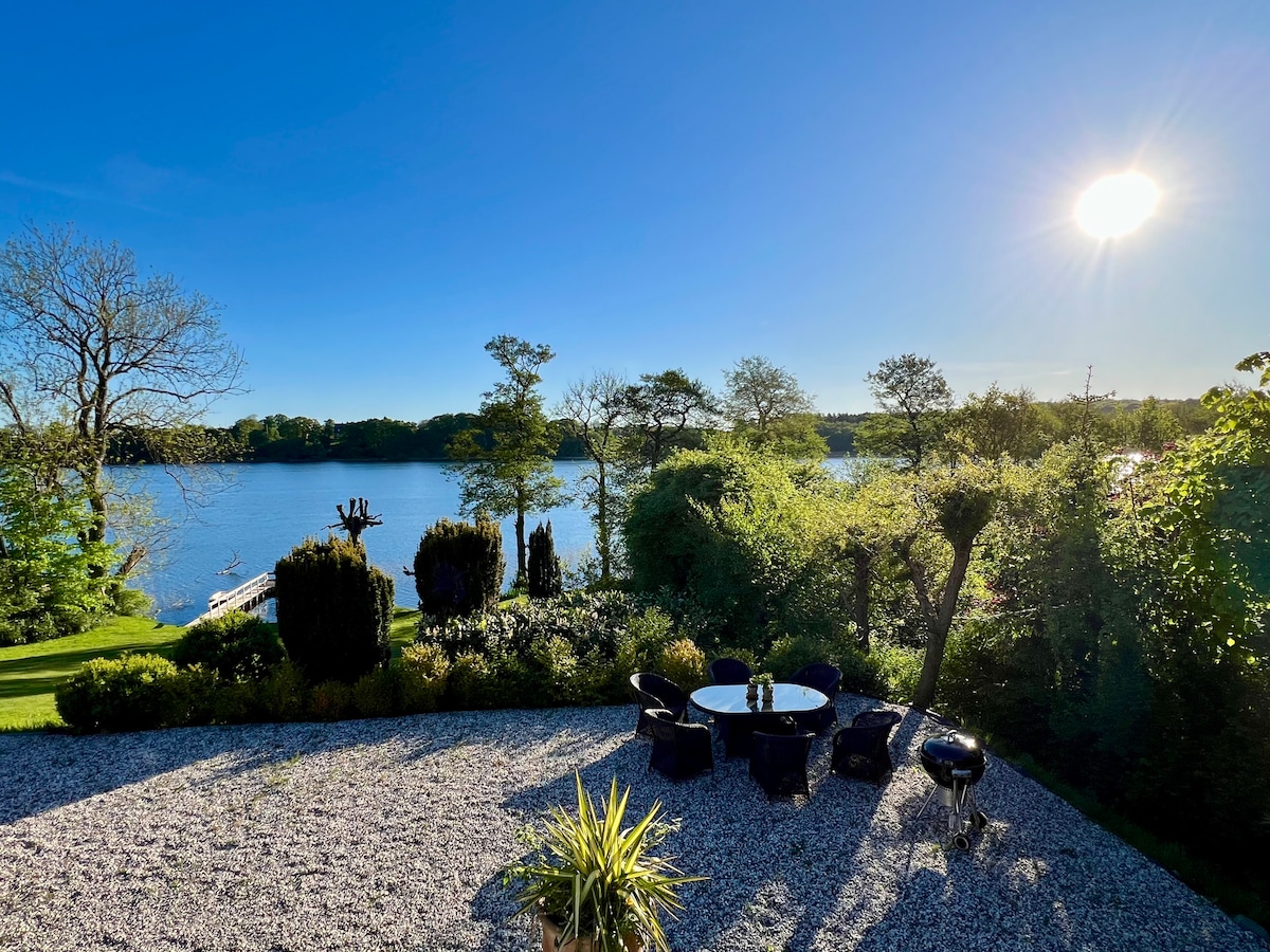 A tranquil outdoor area is set with a circular table and chairs, surrounded by lush greenery and colorful plants. The view encompasses a serene lake shimmering under bright sunlight, with trees lining the opposite shore, promoting a peaceful atmosphere.