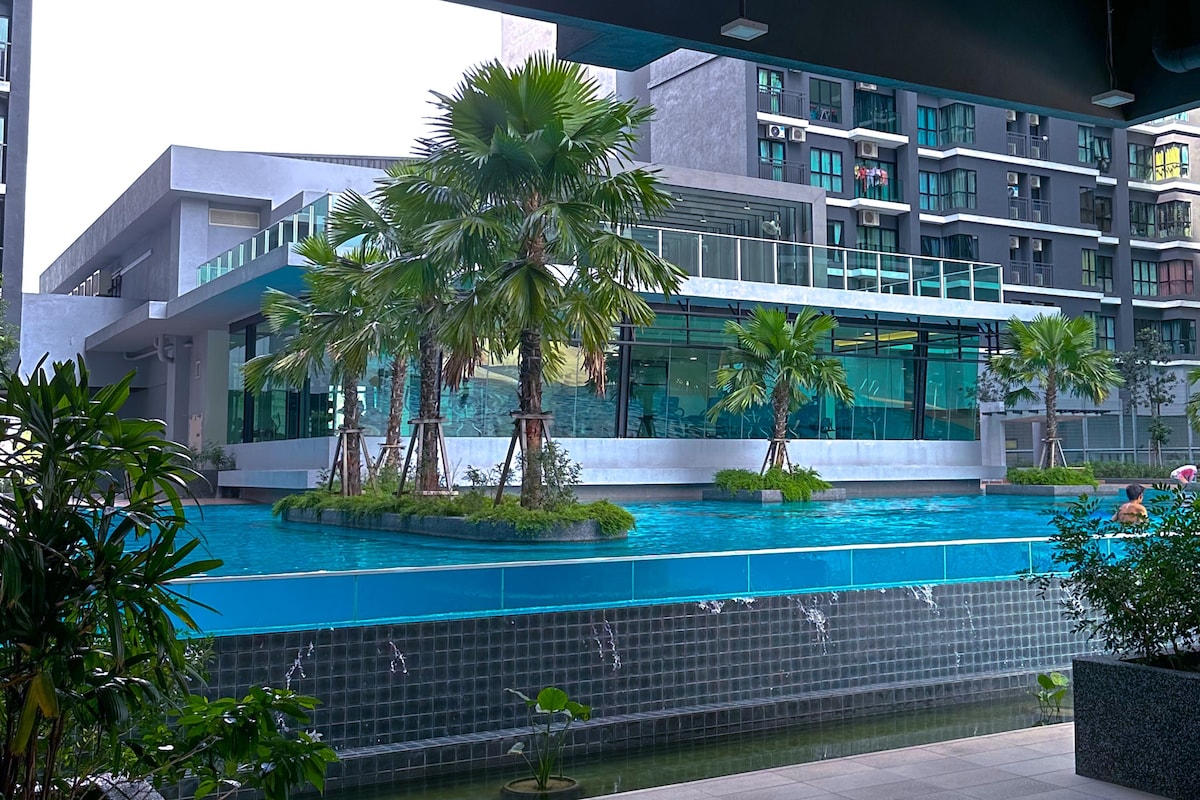 A serene swimming pool is surrounded by landscaped greenery and palm trees. The pool's clear water reflects the modern building facade, which features large windows and outdoor seating areas. Water gently cascades from a wall, creating a tranquil ambiance.
