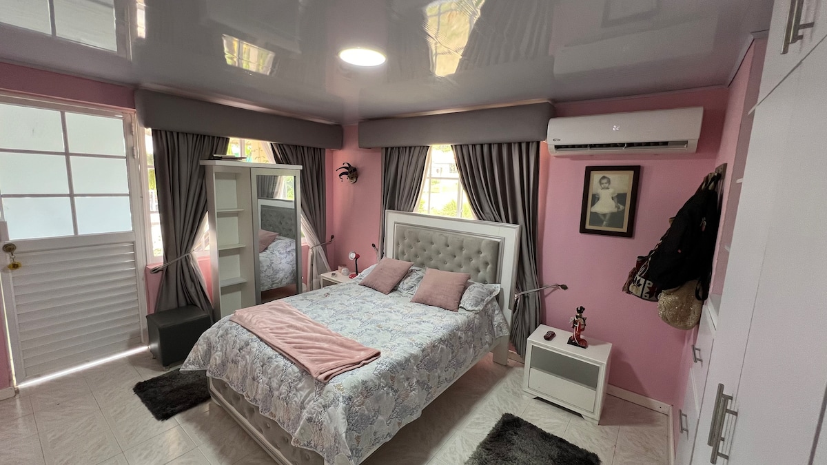 A bedroom with a pink color scheme is shown, featuring a spacious bed dressed in light blue bedding and two pink pillows. Mirrors reflect the room's layout, while gray curtains frame the windows. A nightstand and wardrobe are visible, adding functionality to the space.