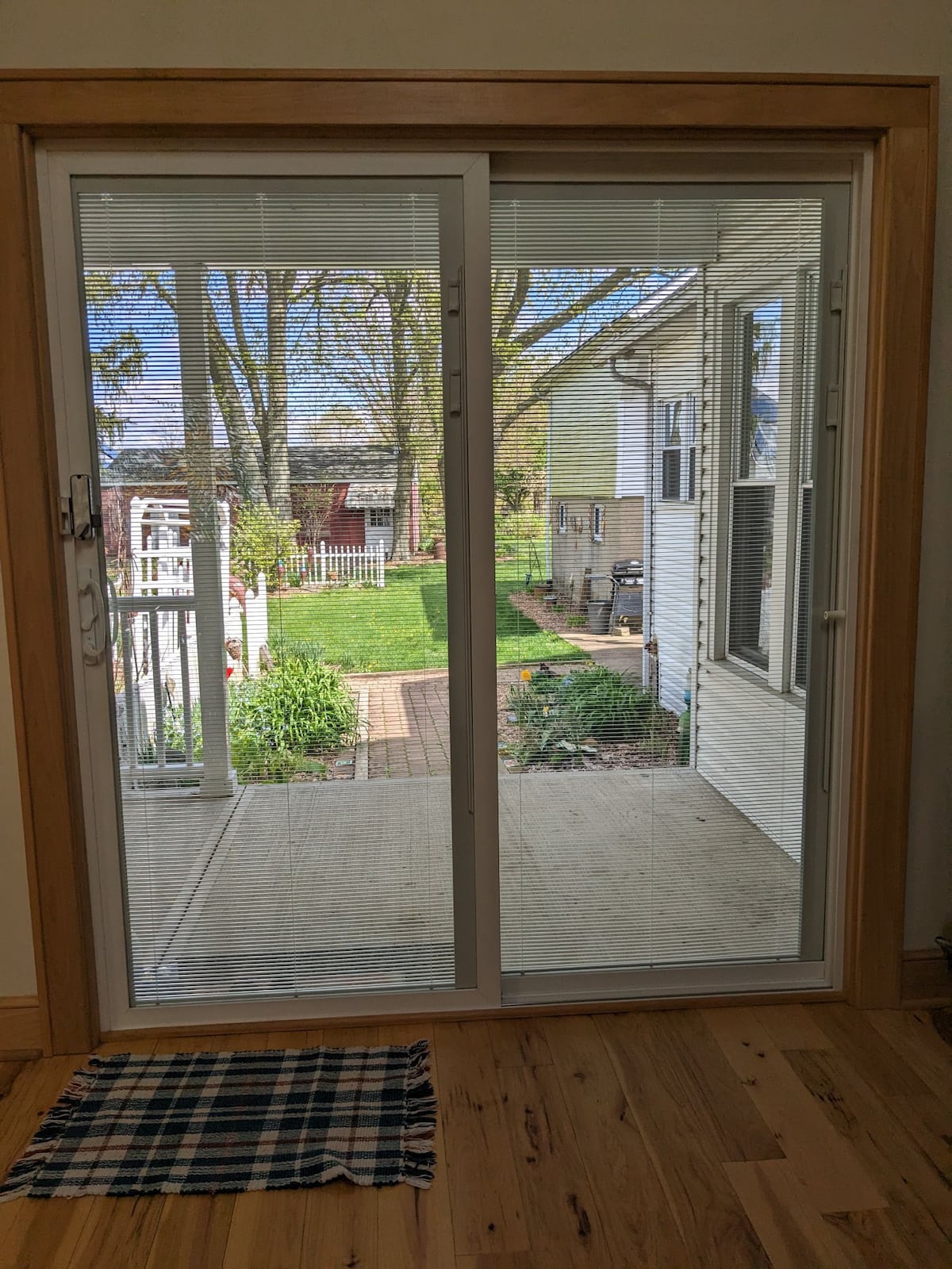 A sliding glass door opens to a view of a green yard, featuring a pathway lined with plants. Sunlight filters through the trees, illuminating the outdoor space. A small welcome mat is placed at the base of the door, adding a homey touch.