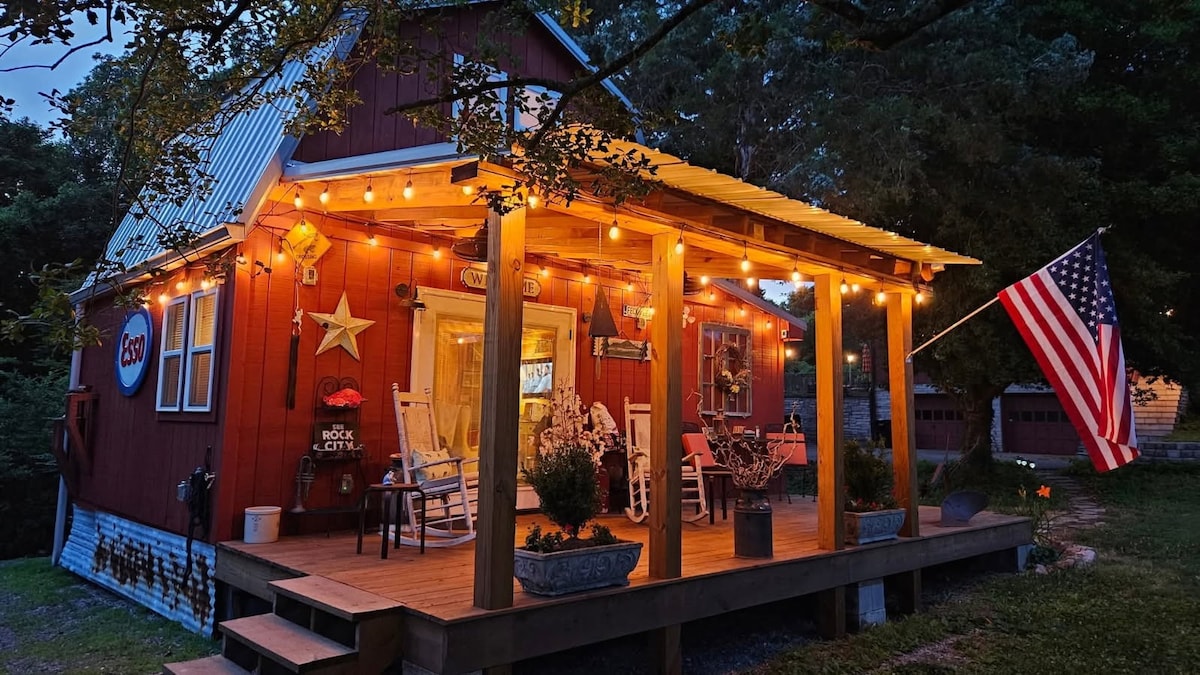 The exterior of the barn features a covered porch adorned with string lights and potted plants. An American flag is displayed prominently. The rustic red barn is complemented by white trim, creating a charming appearance amidst surrounding trees.