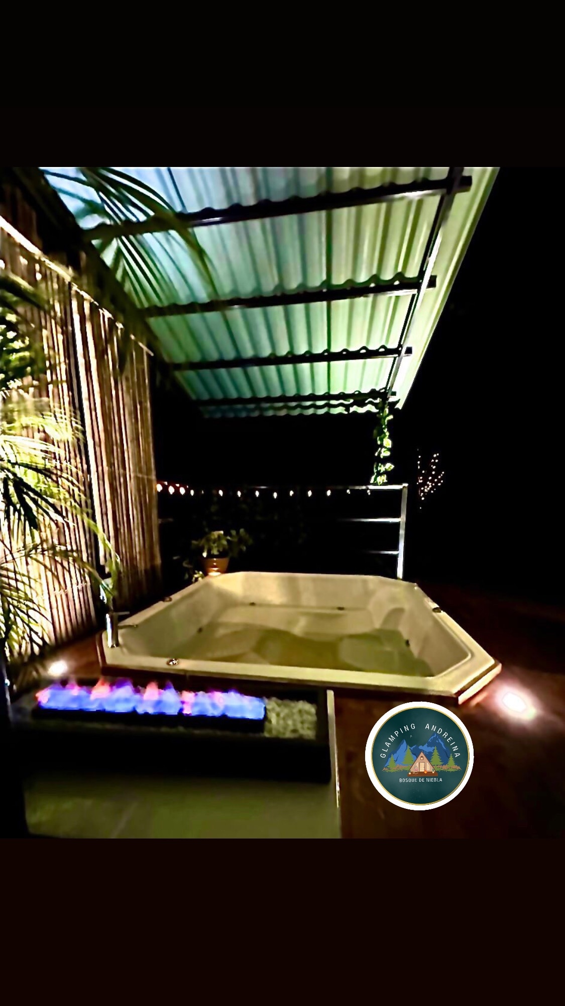 A private hot tub is positioned on a wooden deck, accompanied by a stylish fire feature. Soft lighting from string lights highlights the surrounding greenery, creating a relaxing ambiance. A covered area offers shelter, allowing enjoyment of the space regardless of weather conditions.