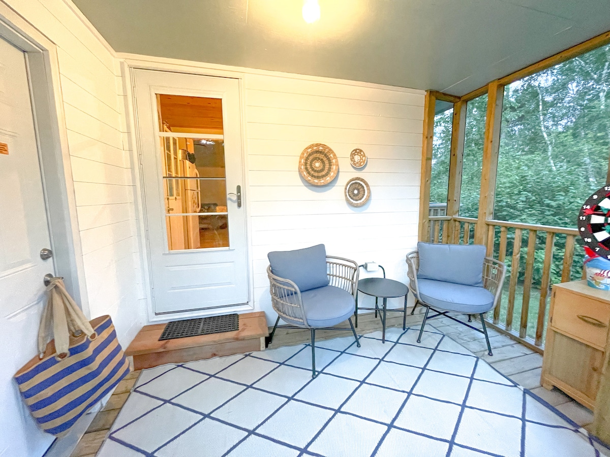 A screened porch features two light blue armchairs paired with a small round table, situated on a patterned rug. The entrance is framed by white wooden walls, with a door leading to the interior. Natural greenery is visible through the screens, enhancing the space's connection to nature.