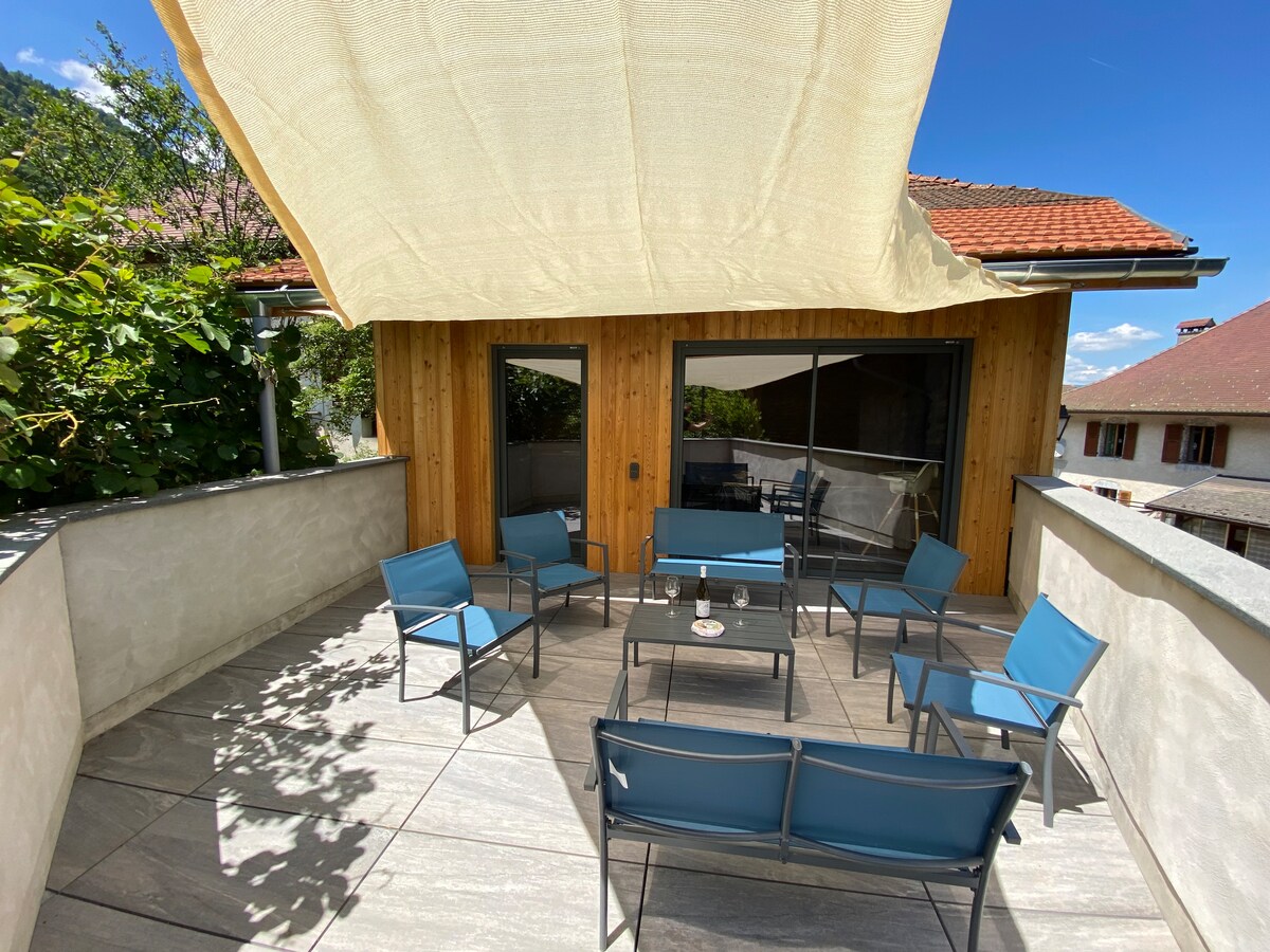 An outdoor terrace is featured with several blue chairs arranged around a central table. A large sunshade provides cover from the sun, while greenery surrounds the space. Large glass doors lead into the interior, inviting natural light into the area.