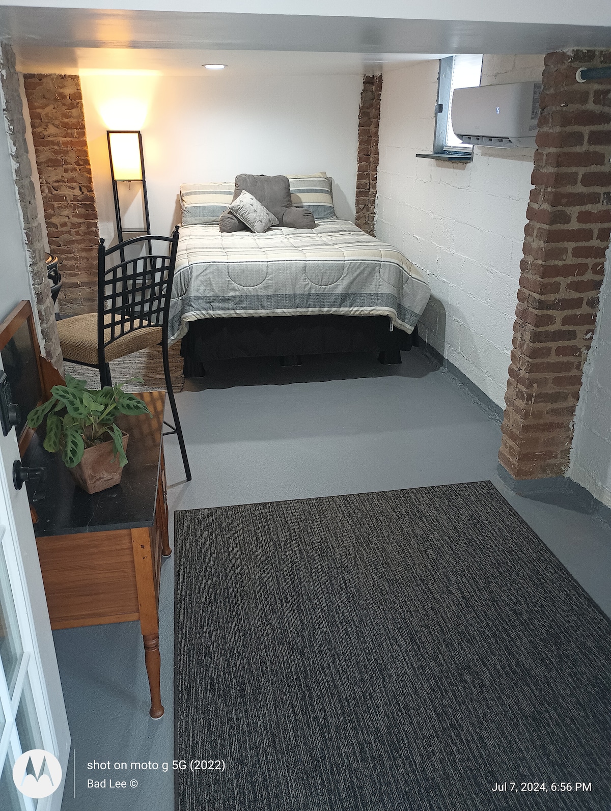A private studio space features a cozy queen bed with a light grey and dark striped comforter. Exposed brick walls add character, while a chair beside the bed offers seating. Natural light filters in through a small window, enhancing the simple decor.