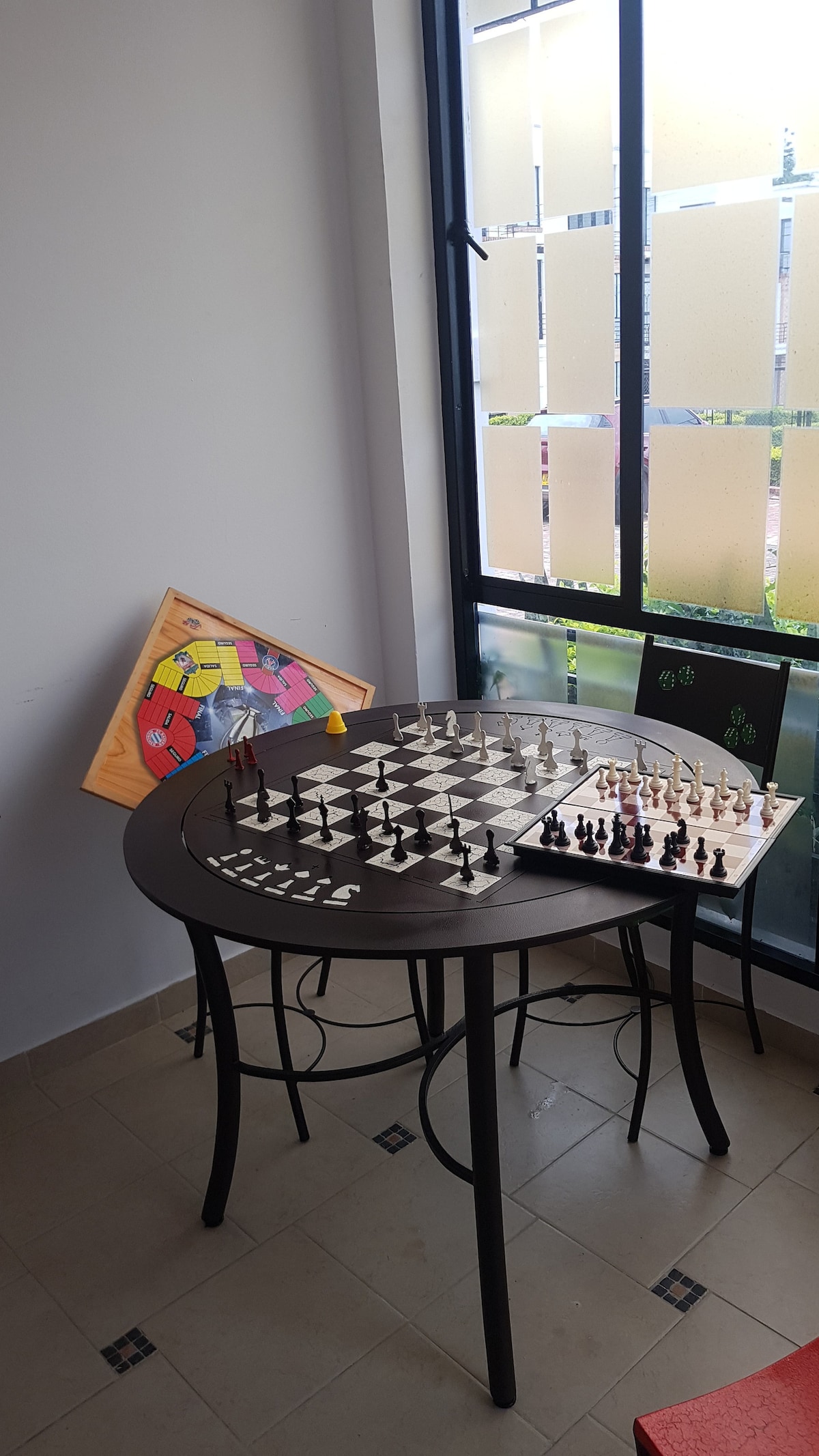 A small table is set with a chessboard displaying pieces in preparation for a game. Nearby, another board game is partially visible against the wall. Natural light filters through large windows, illuminating the gaming area.