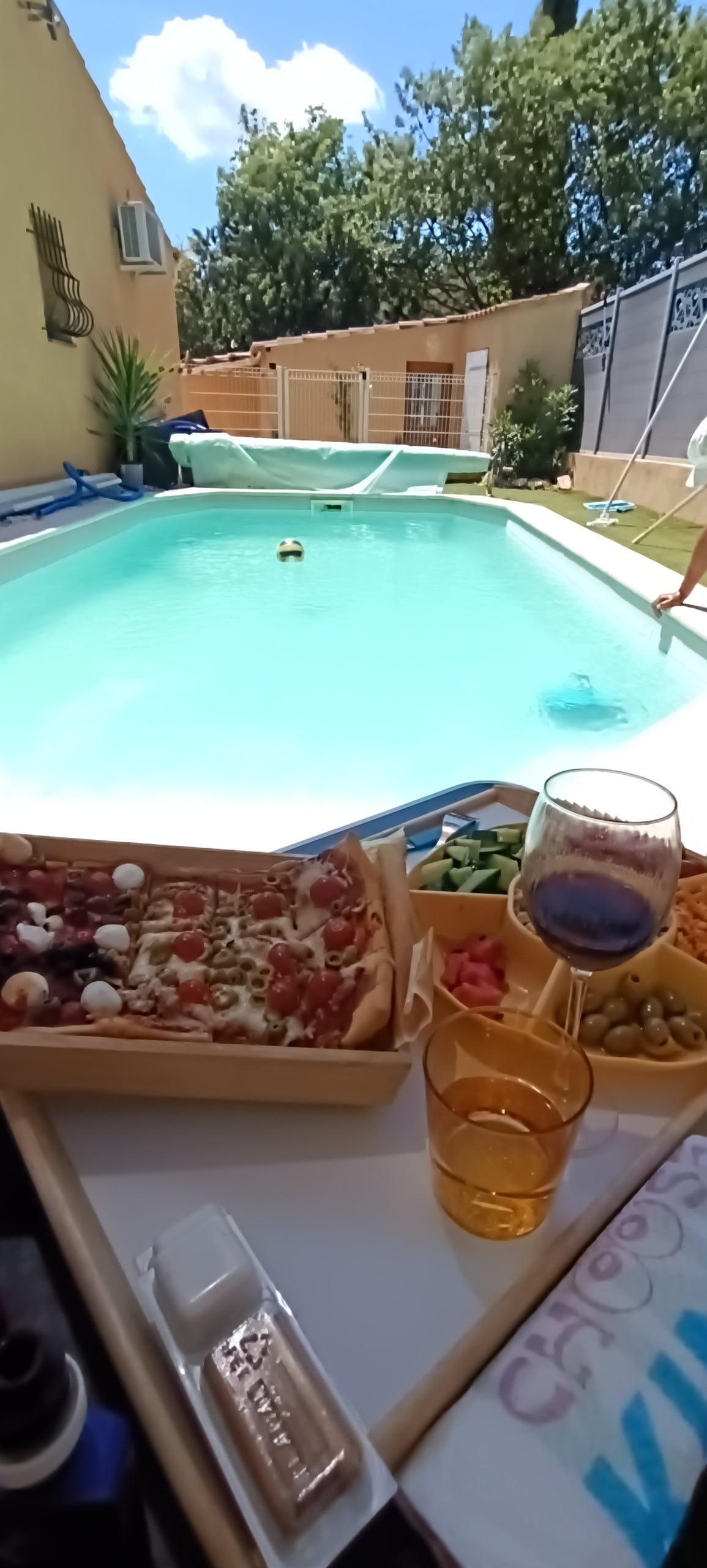 A refreshing pool area is visible, surrounded by neatly arranged fencing. A table is set nearby with a pizza, salad, and two glasses. Sunlight reflects off the water, creating a lively atmosphere. A person can be seen tending to the pool in the background.