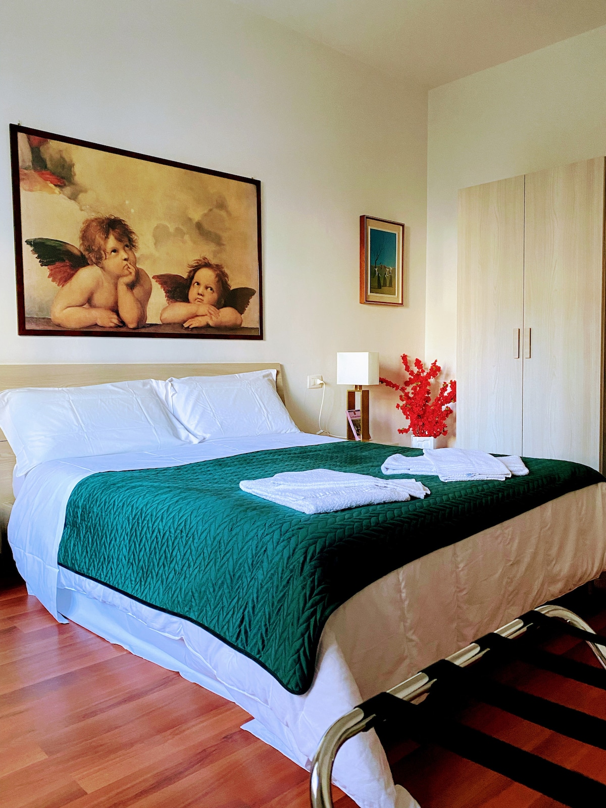 A well-appointed bedroom is visible, featuring a spacious bed covered with a green quilt. Fresh towels are neatly arranged. A framed artwork depicting cherubs adorns the wall, while a small decorative red plant adds a pop of color. A wardrobe is situated to the side.