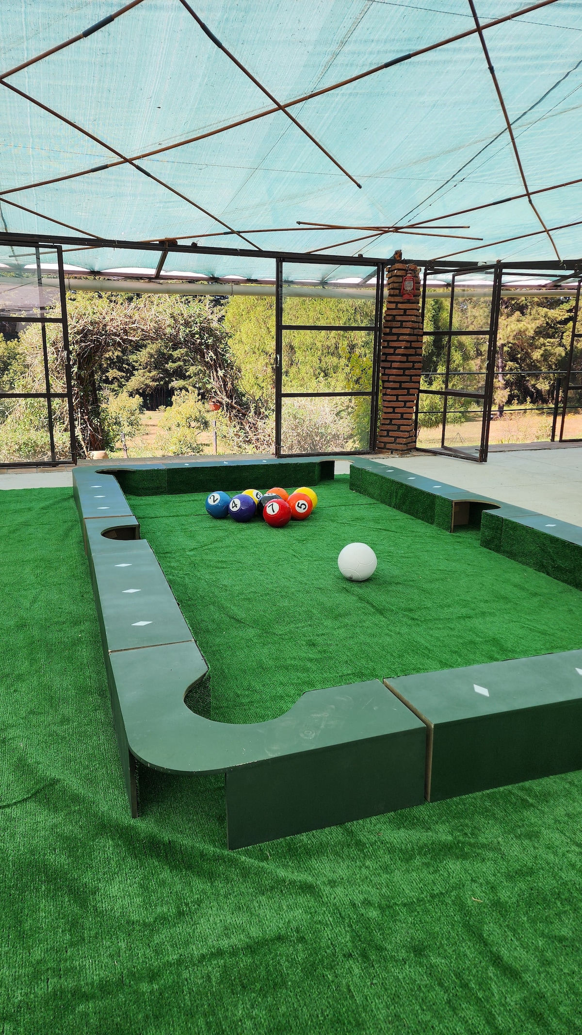 A recreational area is presented with a green, artificial turf surface and a mini-billiards setup. Six colorful balls are arranged on the turf, and a white ball is positioned nearby. The space is enclosed by large glass panels, allowing natural light to fill the area.
