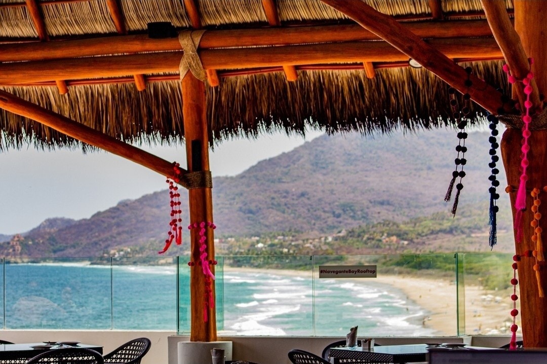 A coastal view showcases rolling waves lapping against a sandy beach, framed by a thatched roof structure. Colorful textiles hang from overhead beams, adding a touch of detail to the spacious terrace, which offers an unobstructed backdrop of mountainous terrain and lush greenery.