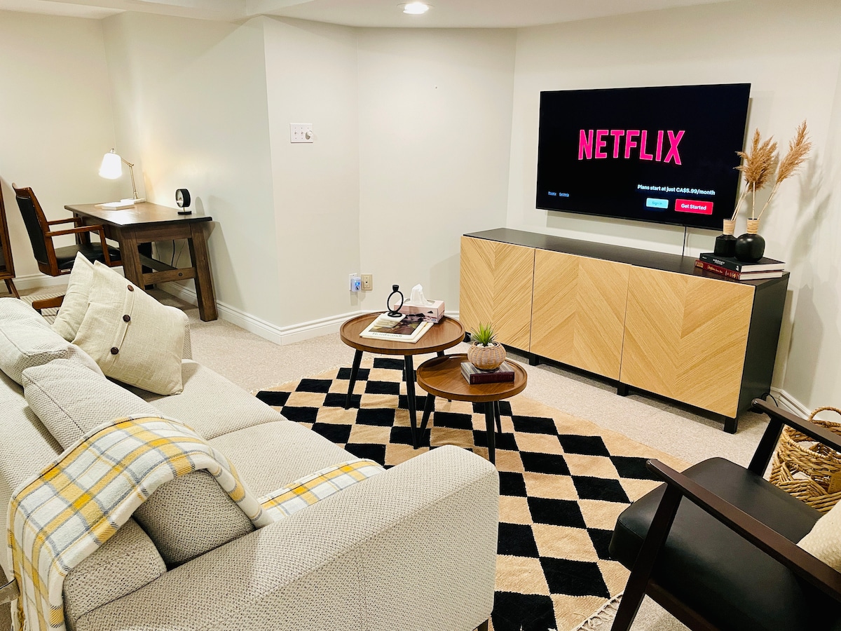 A cozy living area features a neutral-colored sofa adorned with a checkered throw blanket. A stylish coffee table sits on a patterned rug, accompanied by a side chair. A wall-mounted television displays a streaming service, while a light-colored cabinet offers storage and decorative touches.