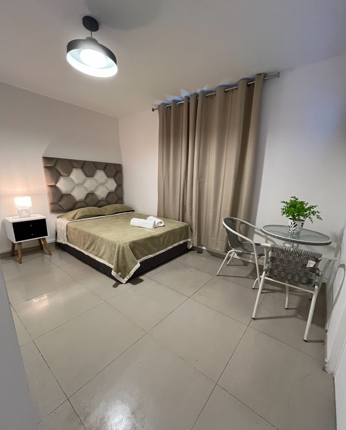 A cozy room features a spacious bed with a neutral-toned headboard and soft bedding. A small dining table with two chairs is positioned nearby, complemented by a vase of greenery. Natural light is partially filtered through floor-length curtains, and a ceiling light provides additional illumination.