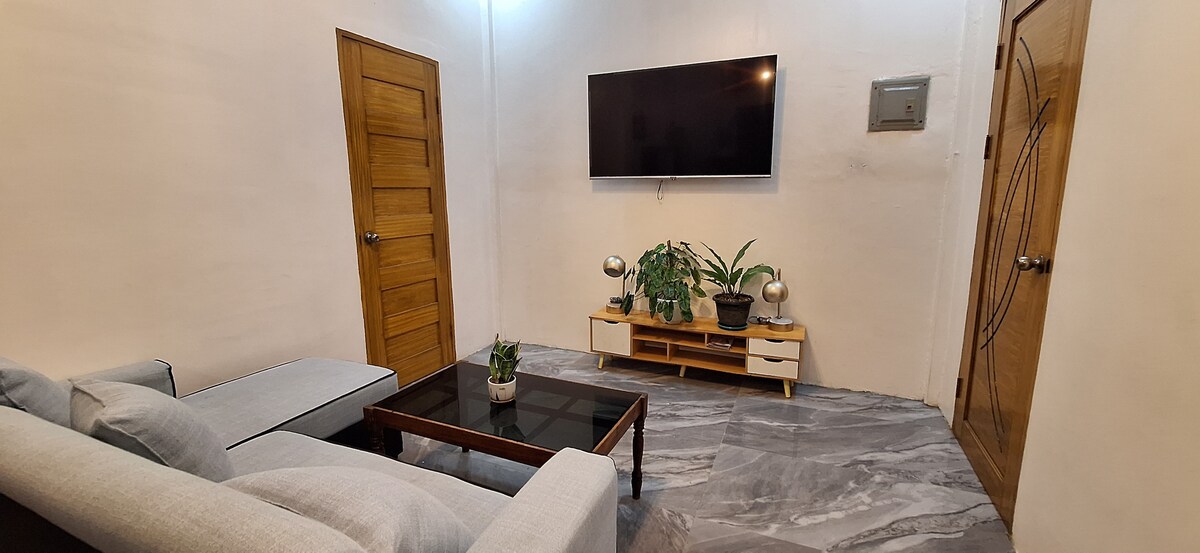 A modern living area is featured with a light gray sofa positioned beside a dark wooden coffee table. A television is mounted on the white wall, above a low-profile cabinet with potted plants, contributing to a minimalist aesthetic.