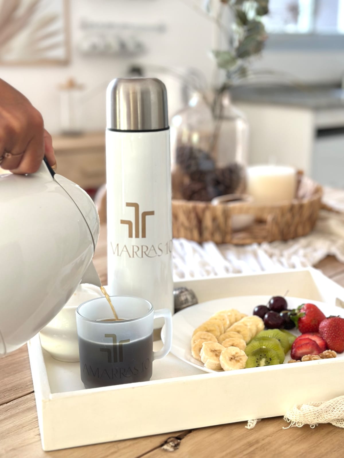 A welcoming breakfast tray is presented with a thermos, a mug, and a plate of fresh fruit, including strawberries, bananas, grapes, and kiwi. Light spills softly across the wooden table, enhancing the inviting setup.