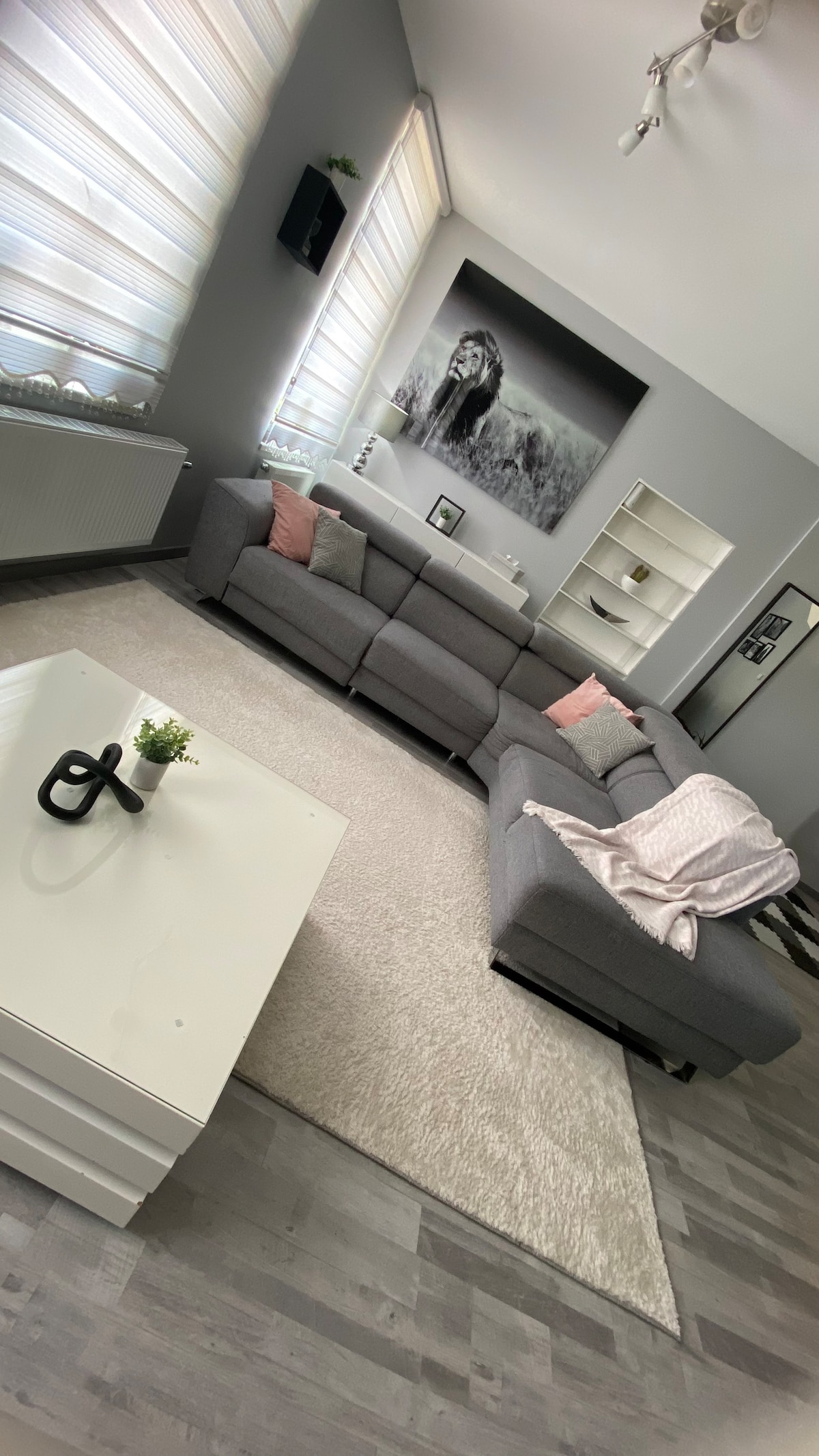 A spacious living room is characterized by a gray sectional sofa adorned with soft pink pillows and a light throw. A white coffee table stands on a plush rug, while a large wall art piece adds visual interest. Natural light filters through layered window treatments.