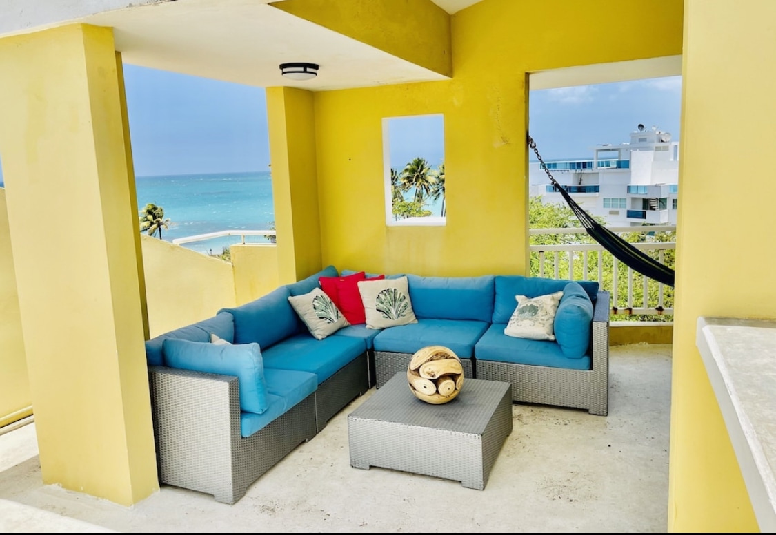 An inviting outdoor lounge area is furnished with a large sectional sofa, accented with colorful pillows. A coffee table sits in the center, complemented by a scenic view of the ocean visible through a nearby window. A hammock is partially visible, enhancing relaxation.